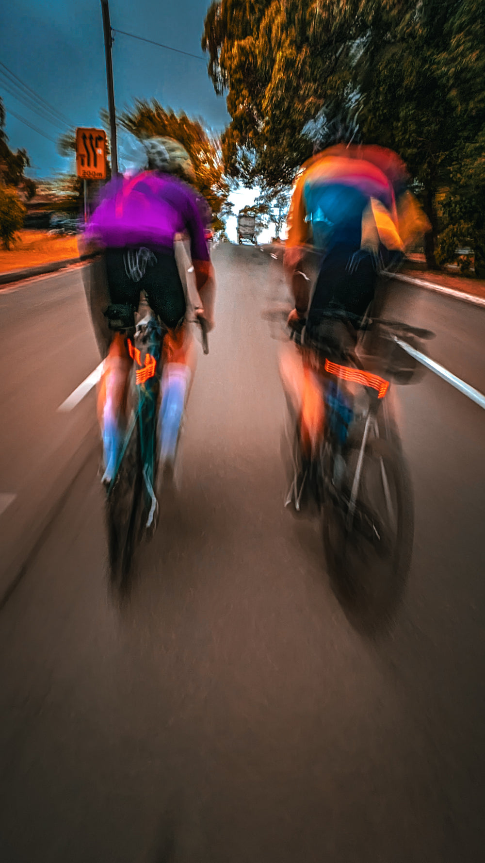 Two cyclists wearing sustainable, premium Aero-Fit cycling clothing ride side by side, their motion creating a blur effect in the background as they move at a high speed. The clothing is designed to improve performance and reduce environmental impact
