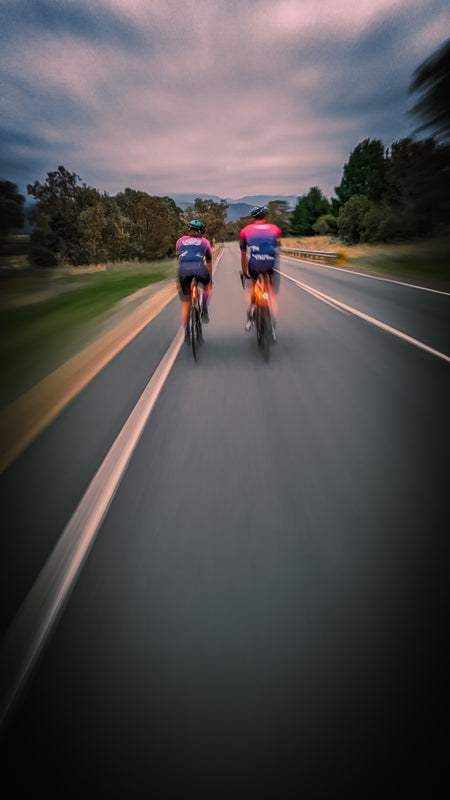 male and female cyclists wearing sustainable, premium Aero-Fit cycling clothing ride side by side through the streets of Canberra, their motion creating a blur effect in the background as they move at a high speed. The clothing is designed to improve performance and reduce environmental impact, while being perfect for the cycling conditions in Canberra