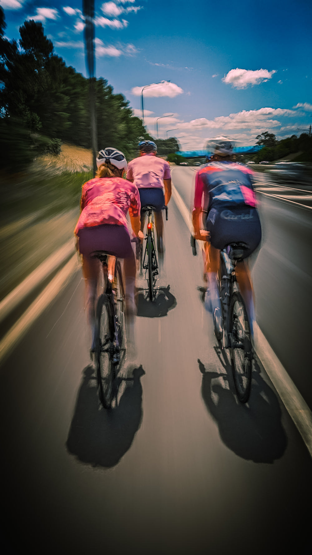 Four cyclists, both male and female, wearing sustainable, premium Aero-Fit cycling clothing ride in a group, their motion creating a blur effect in the background as they move at a high speed. The clothing is designed to improve performance and reduce environmental impact, while providing comfort and style to all genders